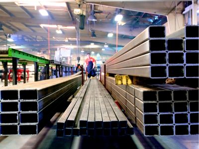 Rows of stacked rectangular and square American-sourced steel tubes and beams stretch into the background of a manufacturing facility, with a person visible in the distance.